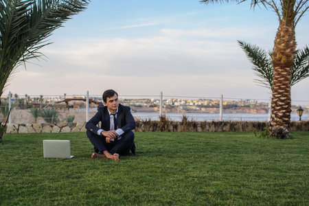 close up photo of a young man in suit with laptop working on green grass near a resort on the...