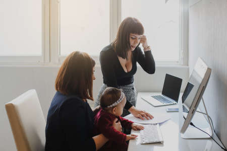 close up photo of two women in office outfit working on computers and discussing a report on the floor; one of them is with a baby girl in her armsの写真素材