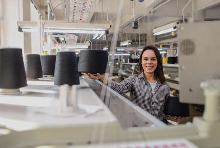 close up photo of a young woman with black threads in cones in her hands near industrial knitting machinesの写真素材