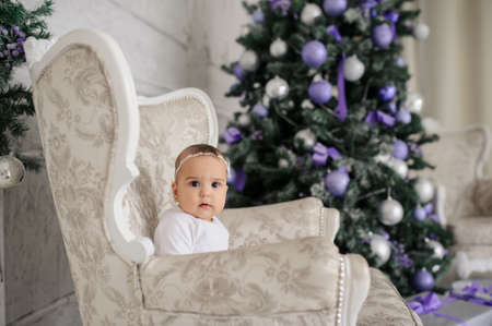 Little baby girl is sitting on a vintage armchair in a white room beautiful decorated for christmas holiday with a Christmas tree and christmas presents.の写真素材