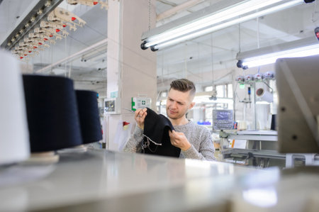 a young man analyzing a knitted piece of cloths near industrial knitted machines with black thread in conesの写真素材