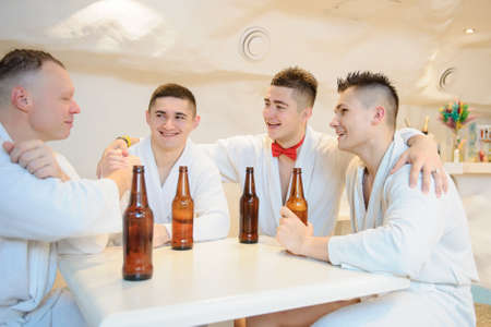 close up photo of 4 men in white gowns sitting around a table and drinking beer after sauna proceduresの写真素材