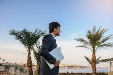 lateral view of a young man in suit with laptop working on green grass near a resort on the seasideの写真素材