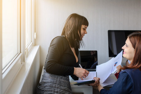lateral view of two women in office outfit working on computers and discussing a reportの写真素材
