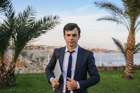 close up photo of a young man in suit with laptop working on green grass near a resort on the...