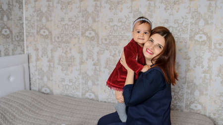 close up photo of mother holding in her arms a one year baby girl in velvet burgundy dress and a grey headband, sitting on a grey blanket on the bed in child's roomの写真素材