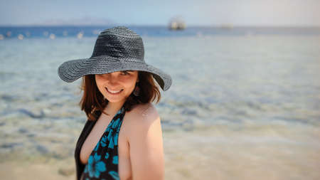 close up photo of a young woman in swimsuit and a blue big hat smiling and sunbathing on the beachの写真素材