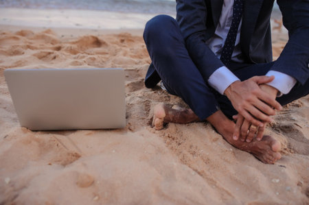 lateral view of a young man in suit with laptop sitting on the beach working on the seasideの写真素材