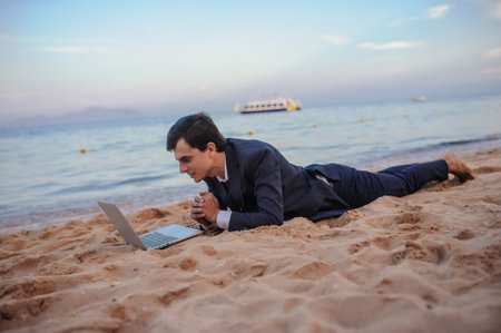 close up photo of a young man in suit with laptop lies on the beach and talking to someone on computerの写真素材