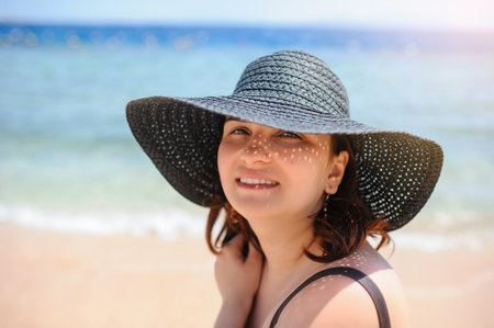 close up photo of a young woman in swimsuit and a blue big hat smiling and sunbathing on the beachの写真素材