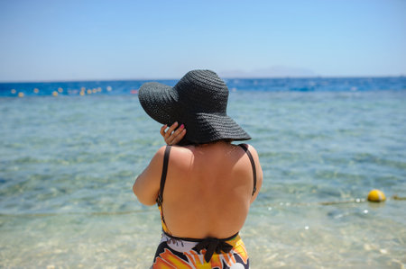 back view of a young woman in swimsuit and a blue big hat sunbathing on the beachの写真素材