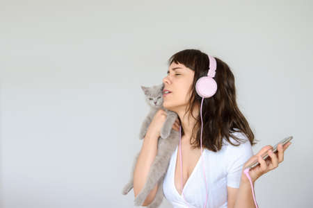 close up photo of a happy brunette with pink headphones listening to the music while holding a gray kitten in her armsの写真素材