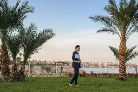 lateral view of a young man in suit with laptop working on green grass near a resort on the seasideの写真素材