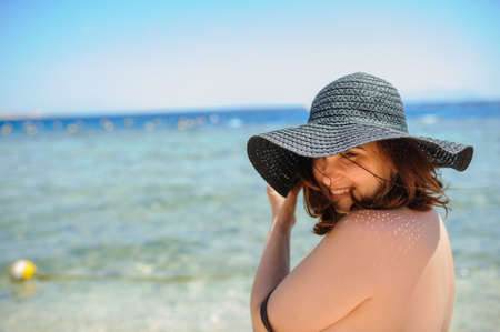 close up photo of a young woman in swimsuit and a blue big hat smiling and sunbathing on the beachの写真素材