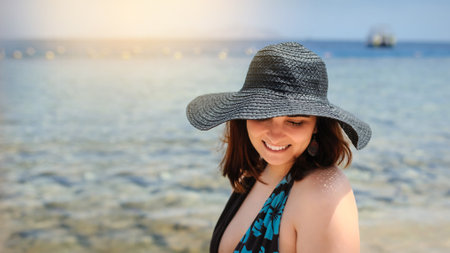 close up photo of a young woman in swimsuit and a blue big hat smiling and sunbathing on the beachの写真素材