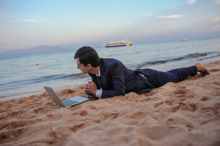 lateral view of a young man in suit with laptop lies on the beach working on the seasideの写真素材