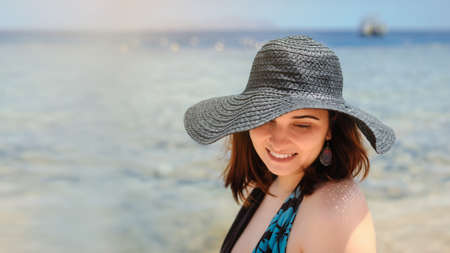 close up photo of a young woman in swimsuit and a blue big hat smiling and sunbathing on the beachの写真素材