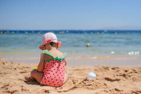 back view photo of a little girl in swimsuit and pink hat sitting on the beachの写真素材
