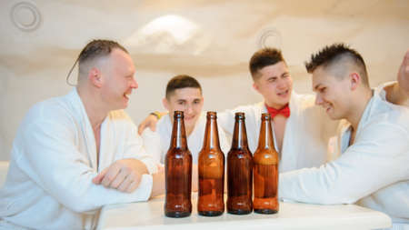 close up photo of 4 men in white gowns sitting around a table and drinking beer after sauna proceduresの写真素材