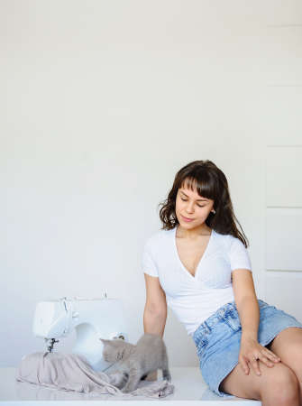 close up photo of a sexy brunette sitting near a white sewing machine and playing with a gray kittenの写真素材