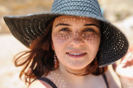 close up photo of a young woman in swimsuit and a blue big hat smiling and sunbathing on the beachの写真素材