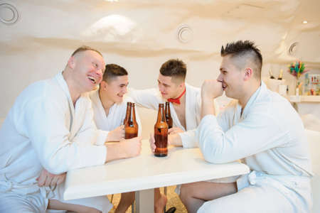 close up photo of 4 men in white gowns sitting around a table and drinking beer after sauna proceduresの写真素材