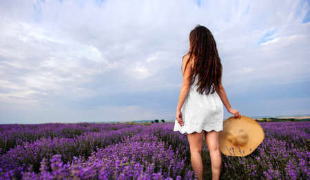 Back view of a young woman in white dress and straw hat walking in the lavender field. Lavender flowers in bloom.の写真素材