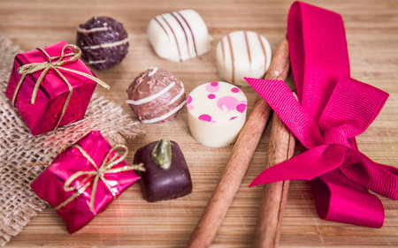 Assorted chocolates with pink ribbon and cinnamon sticks on wooden background. Chocolate pralines...