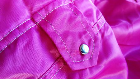 A fragment of a product from a pink raincoat fabric. Sewn white thread pocket with a metallic shiny button. Demi-season women's clothing. Bright glossy synthetic sewing fabric materialの写真素材