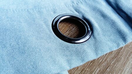 Metal grommet on a gray-blue velvet curtain. The fabric lies on a wooden table. The hole on the material is made of silver coated aluminum. Close-up of a metal eyelet.の写真素材