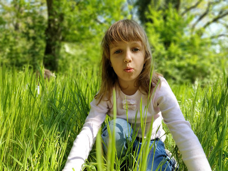 Nice girl in the green grass. The child has blond long hair and a white blouse. The girl looks at...