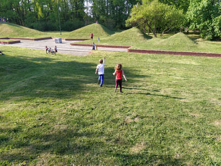 Children run in the park. A boy in a white T-shirt and blue trousers. Girl in a red tank top and burgundy trousers. Green lawn or field. Mowed lawn. People walk in a city park.のeditorial素材