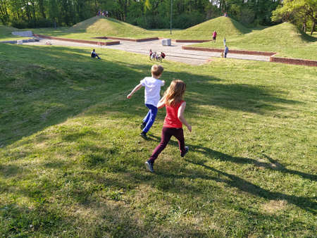 Children run in the park. A boy in a white T-shirt and blue trousers. Girl in a red tank top and burgundy trousers. Green lawn or field. Mowed lawn. People walk in a city park.のeditorial素材