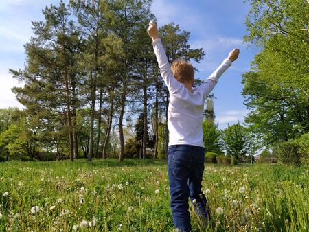 Blond boy stands on the grass and raises his hands up. A child runs in the park. A man is wearing a white longsleeve and blue jeans. Snapshot at the back. Concept of ecology, freedom, victory.の写真素材