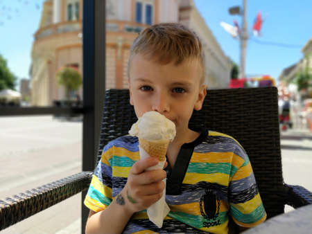 Sremska Mitrovica, Serbia. May 22, 2020 A 7-year-old boy is eating ice cream in a street cafe. A child with a pensive look. In the background, the European city squareのeditorial素材