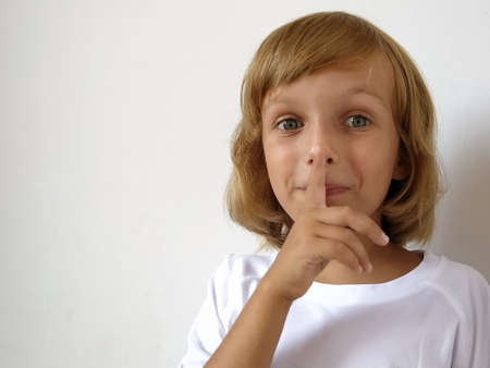 Silence gesture. The finger is near the mouth. The girl on a white background shows a gesture of keeping silence. Child of 7 years old with blond hair. Caucasian type. Copy space. Place for text.の写真素材