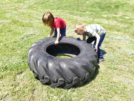 Sremska Mitrovica, Serbia, May 20, 2020. Children play and pick up the tire. Car parts and accessories in a kid's game. A boy and a girl 6 and 7 years old are doing strength exercises on the grass.のeditorial素材