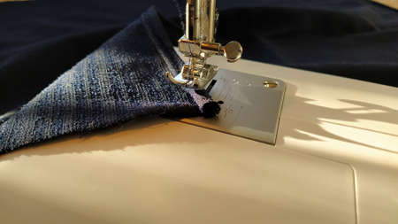 Sewing process on a modern sewing machine, close-up. Sewing machine foot with a needle. Dark blue fabric is trimmed with double wrapping of the fabric edge. Natural sunlight.の写真素材