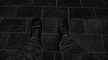 Monochrome black and white image. Feet in boots against the background of street tiles or the road. Two legs of a seated man, photographed from above while sitting.の写真素材