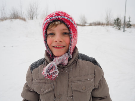 A cute boy 8 years old in a Norwegian knitted red hat with braids and tassels. The child smiles cheerfully. Snowflakes on the face. The theme of winter holidays, frosty weather and travel in the north of Europeの写真素材
