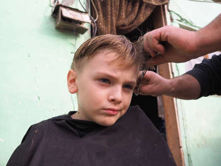 A blond boy of 8 years old gets his hair cut with scissors. Hairdresser services at home. Haircut for a child with blond hairの写真素材