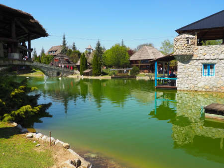 Stanisici, Bijelina, Republika Srpska, Bosnia and Herzegovina April 25, 2021 Ethno village, tourism and attractions. Stone and wood buildings and a bridge. Artificial pond with green water.のeditorial素材