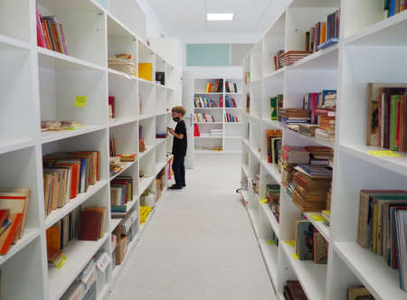 Sremska Mitrovica, Serbia May 15 2021 An 8-year-old boy wearing a mask and a black T-shirt stands near the bookshelf in the library and reads a book. School education. Love for literature and readingのeditorial素材