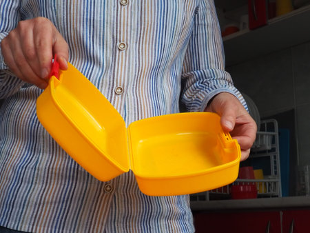 Female hands hold a yellow open plastic school lunch container or packaging. Kitchen utensils. A woman in a white striped shirt shows the lunch box. Food hygiene and recycling theme.の写真素材