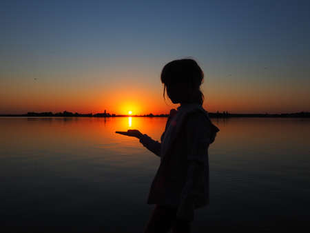 Silhouette of a young girl on a sunset background. The child holds the setting sun on her hand. Horizon, water, sun and sky in the background. Black figure in hooded clothing. Lake Palic, Serbiaの写真素材