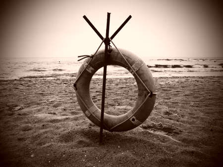 Lifebuoy on a sandy beach. Orange circle on a pole to rescue people drowning in the sea. Rescue point on the shore. Sky and sea in the background. Sepia monochrome. Dark vignetting around the edgesの写真素材