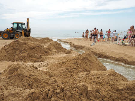 Anapa, Russia, August 19, 2021 A tractor digs a trench on a sandy beach to drain spilled sewage after prolonged downpours. People tourists look at earthworks. Descent of water into the seaのeditorial素材