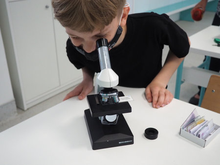 Sremska Mitrovica, Serbia, May 15, 2021. A boy of 8 years old in a black T-shirt looks with one eye through a microscope. Biology lesson. The process of teaching children. School lessons.のeditorial素材