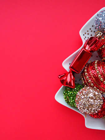 Multicolored Christmas balls and candies on a white plate in the shape of a Christmas tree. Half of the tree is on the right. Copy space Red background. Beautiful striped New Year decor and sparkles.の写真素材