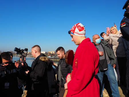 Sremska Mitrovica, Serbia, January 19, 2022. Christian holiday Epiphany. Mass bathing of people in the river in winter. Epiphany bathing. Girl swimmer in a red robe carries a holy cross for ablution.のeditorial素材